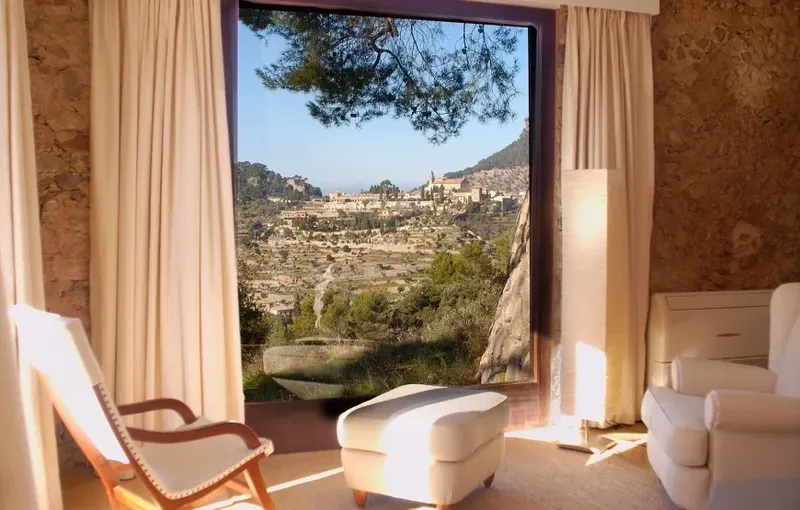 Helles Zimmer mit Sessel und offenem Fenster auf ein Tal mit Dorf am Hang